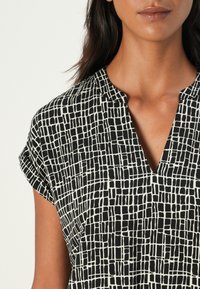Black and white patterned blouse with a V-neckline and rolled short sleeves. Made of lightweight material with a grid texture.