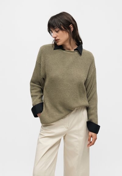 Femme portant un pull en maille vert olive sur une chemise noire à col et un pantalon large crème, debout devant un fond blanc uni.