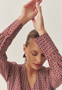 Piros mintás blúz kék részletekkel, hosszú ujjakkal és V-nyakkivágással. Fényes sötét fülbevalók egészítik ki a megjelenést.