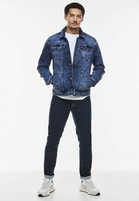 Jeansjacka med en blekt blå tvätt, två bröstfickor, knapplåsning, bärs över en vit skjorta, kombinerad med mörka jeans och vita sneakers.