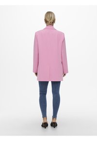 Donna con capelli biondi raccolti in uno chignon, che indossa un blazer rosa, jeans skinny blu e mule nere con tacco, in piedi con la schiena rivolta verso l'osservatore su uno sfondo bianco.