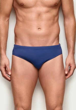 Torso masculino y parte superior de las piernas con bañador tipo slip azul marino, de pie frente a un fondo claro y liso.