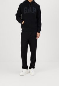 Uomo che indossa una felpa nera GAP con cappuccio, pantaloni neri e sneakers bianche, in piedi contro uno sfondo bianco semplice.
