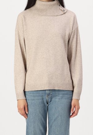 Femme portant un pull à col roulé côtelé beige avec des boutons au col et un jean bleu, debout sur un fond blanc.