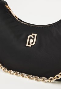 Mustmust kott mustast nailonist, kullatoonsete ketti- ja tõmbelukuga detailidega. Ees on logo aktsent ja tekstuur on sile.