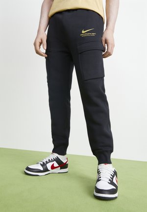 Pantalones de chándal negros con bolsillos de carga, con un logo dorado de Nike y texto atlético. Combinados con zapatillas blancas, acentos rojos y una camiseta amarilla.