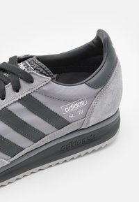 Scarpa atletica grigia con un mix di camoscio e tessuto, tre strisce scure, un tallone nero testurizzato e una suola in gomma nera.