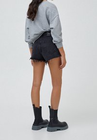 Mujer vestida con pantalones cortos de mezclilla negros deshilachados, sudadera gris y botas de tobillo negras con suela gruesa, de pie frente a un fondo simple.