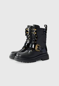 Botas de tobillo de cuero negro con una suela gruesa, acentos brillantes, herrajes dorados y diseño con cordones; cuentan con una hebilla decorativa en el lateral.