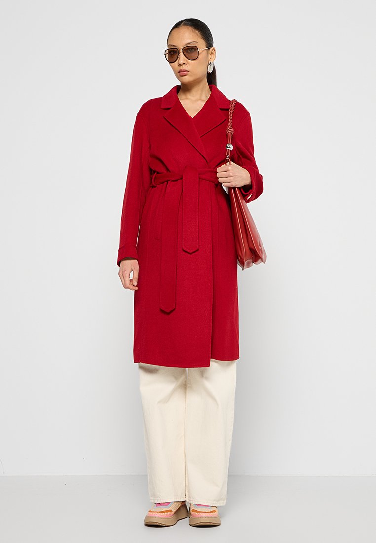 Roter Wollmischung-Trenchcoat mit tailliertem Gürtel, Knopfleiste und Reverskragen, kombiniert mit cremefarbenen weiten Hosen und braunen Schuhen.