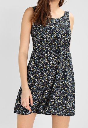 Vrouw draagt mouwloze zwarte minikleding met kleine meerkleurige bloemenprint en oorringen, staand tegen een lichtgrijze achtergrond.