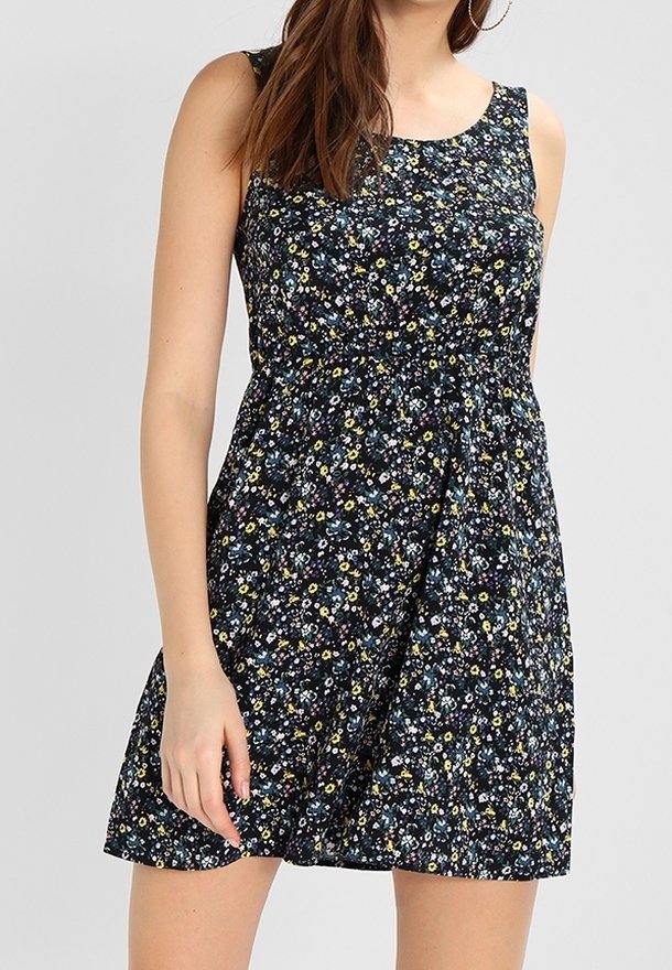 Femme portant une mini-robe noire sans manches avec un petit imprimé floral multicolore et des boucles d’oreilles créoles, debout devant un fond gris clair.