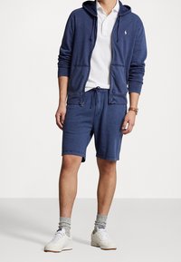 Marinblå zip-up hoodie med dragsko, i kombination med matchande shorts. Har en vit pikétröja underneath och vita sneakers.