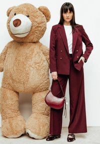 Blazer bordeaux à double boutonnage et pantalon à jambes larges, top blanc, tenant un sac en cuir bordeaux, à côté d'un grand ours en peluche beige.