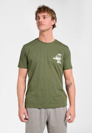 Mand iført en olivengrøn t-shirt med hvidt grafik på brystet, lysegrå bukser og en tatovering på venstre underarm, stående mod en ensfarvet baggrund.