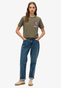 Olivengrøn bomulds t-shirt med et farverigt grafisk design, der viser en svane og blomster; parret med blå denimjeans og cremefarvede sneakers.