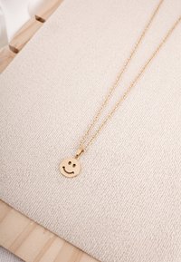 Złoty naszyjnik z zawieszką w kształcie uśmiechniętej buźki, z delikatnym łańcuszkiem na tle teksturowanej beżowej tkaniny.