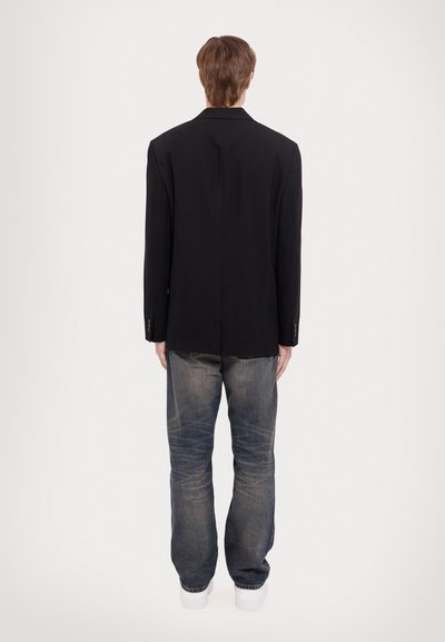 Schwarzes Blazer im geraden Schnitt, kombiniert mit lockeren blauen Jeans. Das Blazer hat einen Reverskragen und zwei Rückenschlitze. Weiße Sneaker runden das Outfit ab.