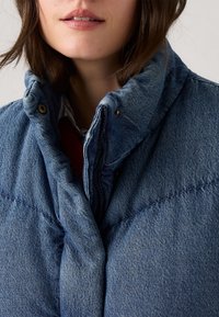 Denim puffervest med høy krage, med quiltet design, trykknapper og en myk tekstur. Dyp blå farge med lag under.