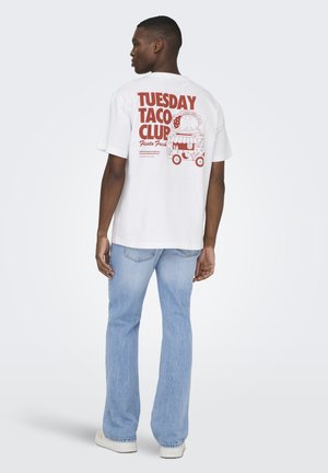 Uomo che indossa una maglietta bianca con scritta rossa "Tuesday Taco Club" e illustrazione di un camioncino taco, abbinata a jeans azzurri e scarpe bianche.