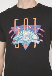 Juodas medvilninis marškinėliai su spalvingu grafikiniu spaudiniu, kuriame yra teksto ir formų, įskaitant mėlyną trikampį ir juostelę su užrašu "Levi Strauss & Co."
