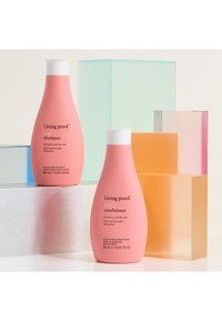Deux bouteilles roses de shampoing et après-shampoing pour boucles Living Proof avec des bouchons blancs, entourées de blocs de verre colorés et géométriques.