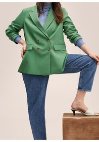 Zielona dwurzędowa marynarka z złotymi guzikami, w połączeniu z jasnoniebieskim golfem, luźnymi niebieskimi dżinsami i białymi mule na obcasie na drewnianym pedałku.