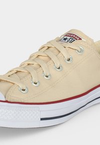 Μπεζ χαμηλού τύπου παντοφλέ Converse από καμβά με λευκή λαστιχένια σόλα, κόκκινη ρίγα, μεταλλικά κρικάκια και μπεζ κορδόνια, εμφανίζοντας μπροστινές και πλευρικές λεπτομέρειες.