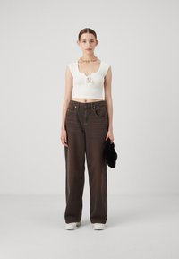 Hvit cropped topp med snøring foran, kombinert med mørkebrune vide jeans. Modellen holder et svart pelslignende tilbehør. Enkel bakgrunn.