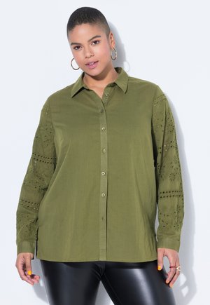 Kobieta w oliwkowej koszuli na guziki z haftowanymi koronkowymi rękawami i czarnych skórzanych spodniach, stojąca na tle jednolitego jasnego tła.