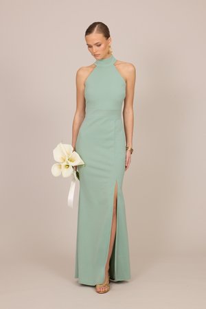 Kobieta w miętowej sukni na szyję z rozcięciem na udzie trzyma białe kalie, nosi złote sandały, kolczyki i bransoletkę na beżowym tle.