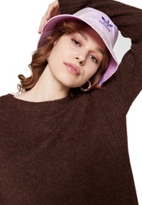 Gaiši violets buktas cepure ar izšūtu tekstūru un logo, apvienota ar brūnu adītu džemperi. Redzama gluda āda un izsmalcināts grims.