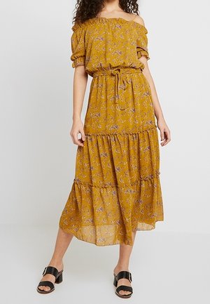Robe moutarde décolletée, avec un motif floral, une taille cintrée, un ourlet à volants et de courtes manches bouffantes. Associée à des sandales noires à bout ouvert.