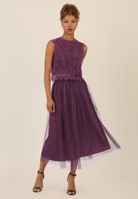 Violetti mekko, jossa on pitsinen yläosa, jossa on aaltoilevat reunat, läpinäkyvä tyllihame, jossa on pilkkuja, ja mustat nilkkaremmikengät.