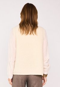 Un pull sans manches de couleur crème avec une finition en tricot texturé, un ourlet côtelé et une coupe légèrement oversize. Porté par-dessus une chemise légère à manches longues.