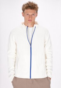 Giacca bianca con zip e colletto alto, caratterizzata da dettagli in zip blu. Tessuto morbido e tasche laterali. Indossata sopra una camicia chiara e pantaloni beige.