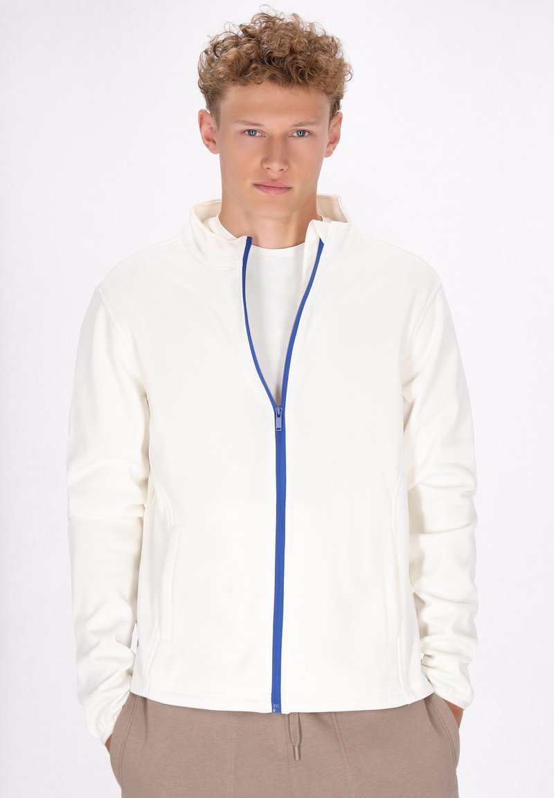 Vit zip-up jacka med hög krage, med blå dragkedja. Mjuk tyg och sidofickor. Bärs över en ljus skjorta och beige byxor.