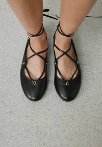 Sorte læderballerinaer med krydsede ankelbind, ovalt tåpanel og metalaksenter. Glat tekstur med minimalistisk design på en grå overflade.