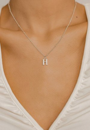 Silberne Halskette mit einem strukturierten Buchstaben "H" Anhänger, vor einem minimalistischen weißen Oberteil mit V-Ausschnitt, das den Schmuck hervorhebt.