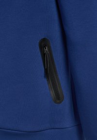 Tessuto blu con tasca verticale nera con cerniera e linguetta nera, sullo sfondo una vista ravvicinata di un capo di abbigliamento.