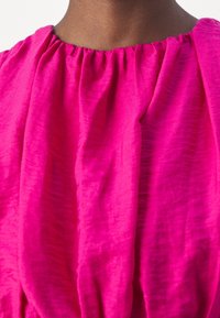 Prenda de color rosa brillante con un escote fruncido. La tela es texturizada, mostrando suaves y sutiles arrugas. Enfoque en el detalle rizado del cuello.