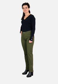 Top negro de manga larga y escote en V combinado con pantalones ceñidos en verde oliva, que presentan acentos de botones plateados en los puños, y botines de tacón negros.