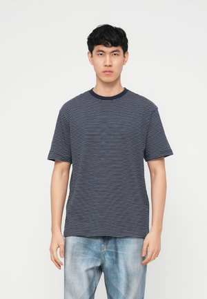 Jonge man met kort zwart haar, gekleed in een marineblauw-wit gestreept T-shirt en lichtblauwe spijkerbroek, staand tegen een effen witte achtergrond.