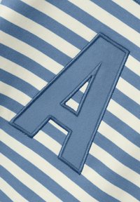 Parcha de tela azul con la forma de la letra "A" cosida sobre un fondo textil blanco y azul a rayas horizontales.