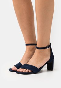 Mörkblå mocka högklackade sandaler med ankelrem och klackfot. Har breda framremmar och öppen tå-design.