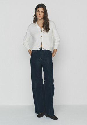 Moterys su ilgais tamsiais plaukais, dėvinčios baltą mygtukais užsegamą megztinį ir tamsius platėjusius džinsus, stovinčias su rankomis kišenėse.