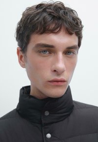 Black puffer jacket with a high collar, featuring silver snap buttons. Close-up of a person’s face with short, curly hair and light blue eyes.