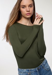Body z długim rękawem w oliwkowym kolorze, wykonany z miękkiego materiału, z dekoltem typu łódka, zestawiony z jasnoniebieskimi dżinsami o wysokim stanie.