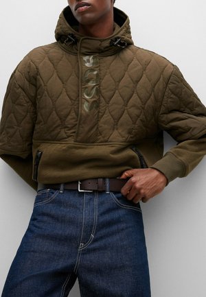 Mann trägt gesteppte olivgrüne Kapuzenjacke mit BOSS-Logo, dunkelblaue Jeans und braunen Gürtel, Hand ruht auf der Gürtelschnalle.