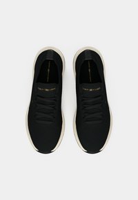 Black knit sneakers with a textured upper, featuring flat laces and a white outsole. Gold logo on the tongue adds contrast.